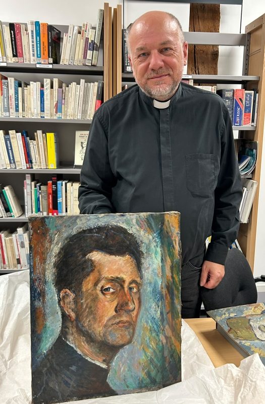 Un homme portant une chemise noire se tenant debout, tenant un tableau représentant un portrait en arrière-plan d'une bibliothèque.